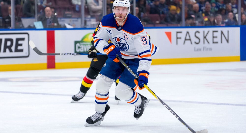 Foto em ação: Connor McDavid, atacante do Edmonton Oilers, com o número 97 no uniforme, manuseia o disco durante partida contra o Vancouver Canucks na Rogers Arena. O jogador, usando uniforme azul escuro, se inclina sobre o gelo; ao fundo, um jogador em uniforme preto tenta interceptar.