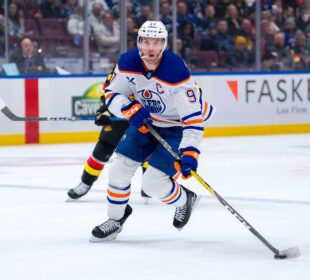 Foto em ação: Connor McDavid, atacante do Edmonton Oilers, com o número 97 no uniforme, manuseia o disco durante partida contra o Vancouver Canucks na Rogers Arena. O jogador, usando uniforme azul escuro, se inclina sobre o gelo; ao fundo, um jogador em uniforme preto tenta interceptar.