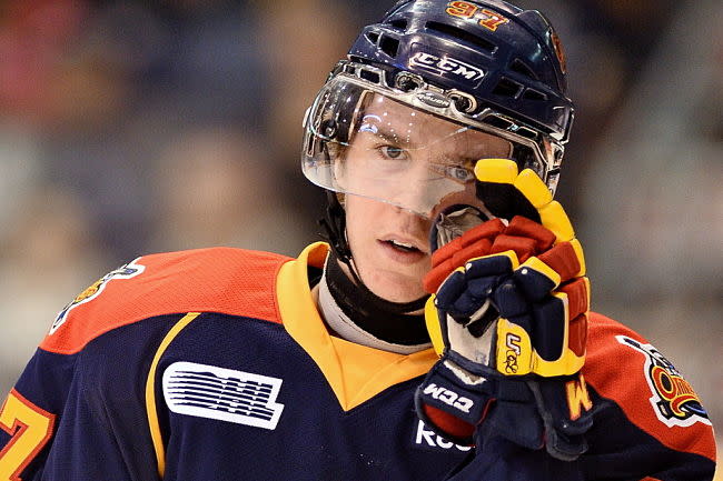 Connor McDavid, aos 17 anos, em ação pelo Erie Otters. Ele veste uniforme azul escuro, laranja e amarelo, com capacete azul e luvas de hóquei,