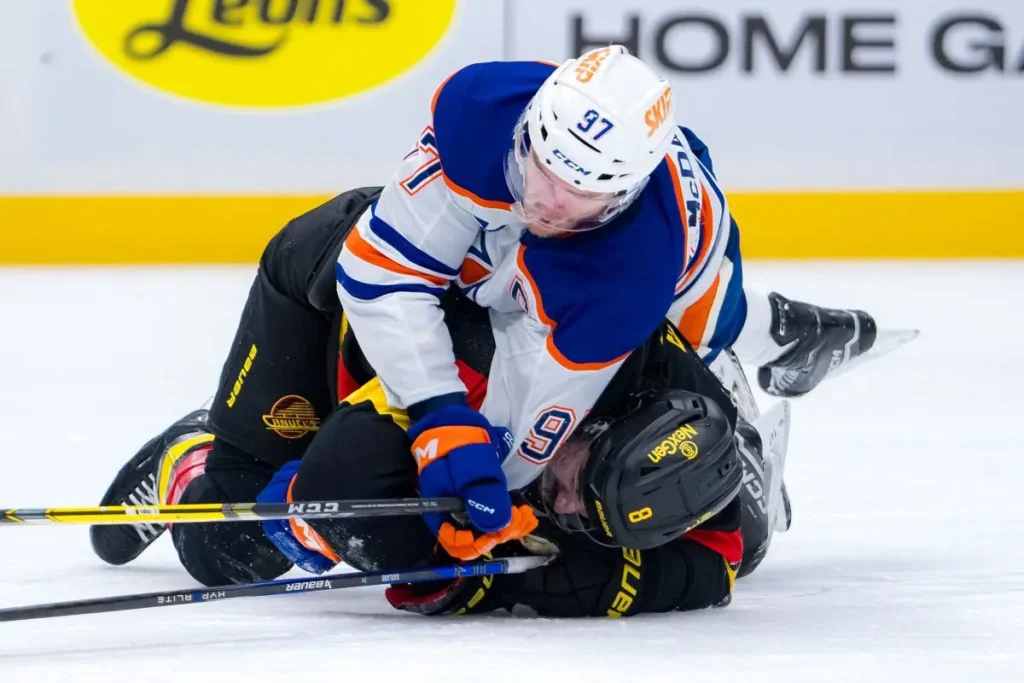 Connor McDavid (#97/Oilers, uniforme branco e azul) está por cima de Conor Garland (#08/Canucks, uniforme preto e amarelo) durante partida. McDavid, em posição dominante, pressiona Garland sobre o gelo, ambos com tacos de hóquei